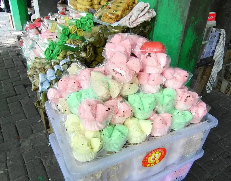 Bolu kukus warna merah muda, hijau, dan kuning dalam plastik ditumpuk di lapak jajanan pasar