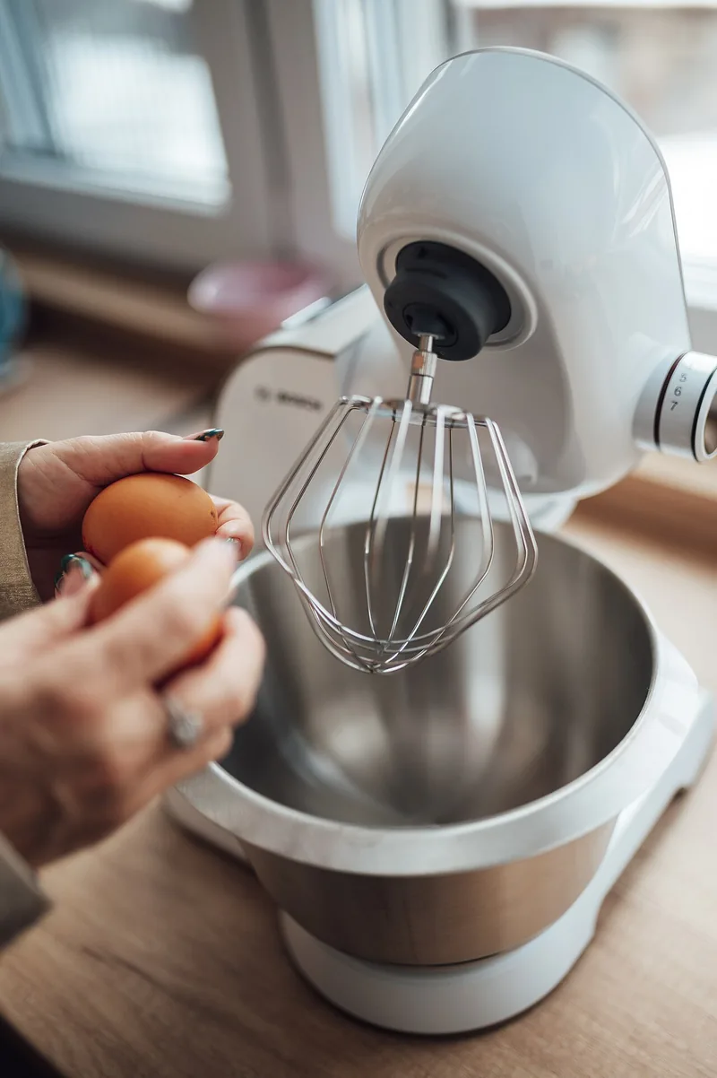 Tangan memegang dua butir telur di samping mangkuk mixer berpengocok balon yang siap dipakai