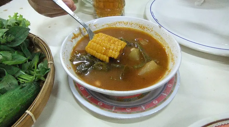 Sayur asem berkuah merah kecokelatan dengan jagung dan labu siam disendok dari mangkuk, lalapan di sampingnya