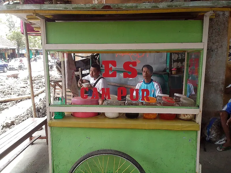 Gerobak hijau penjual es campur di pinggir jalan dengan deretan toples bahan di etalase