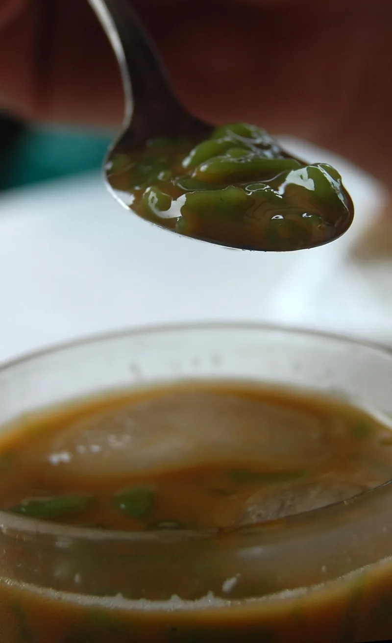 Butiran cendol hijau di sendok dengan mangkuk minuman di bawahnya