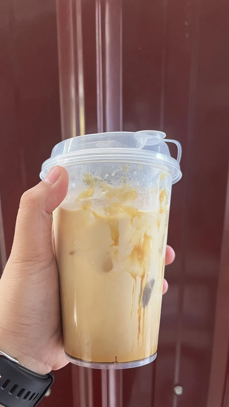 Tangan memegang gelas plastik es kopi susu gula aren berwarna krem dengan es batu