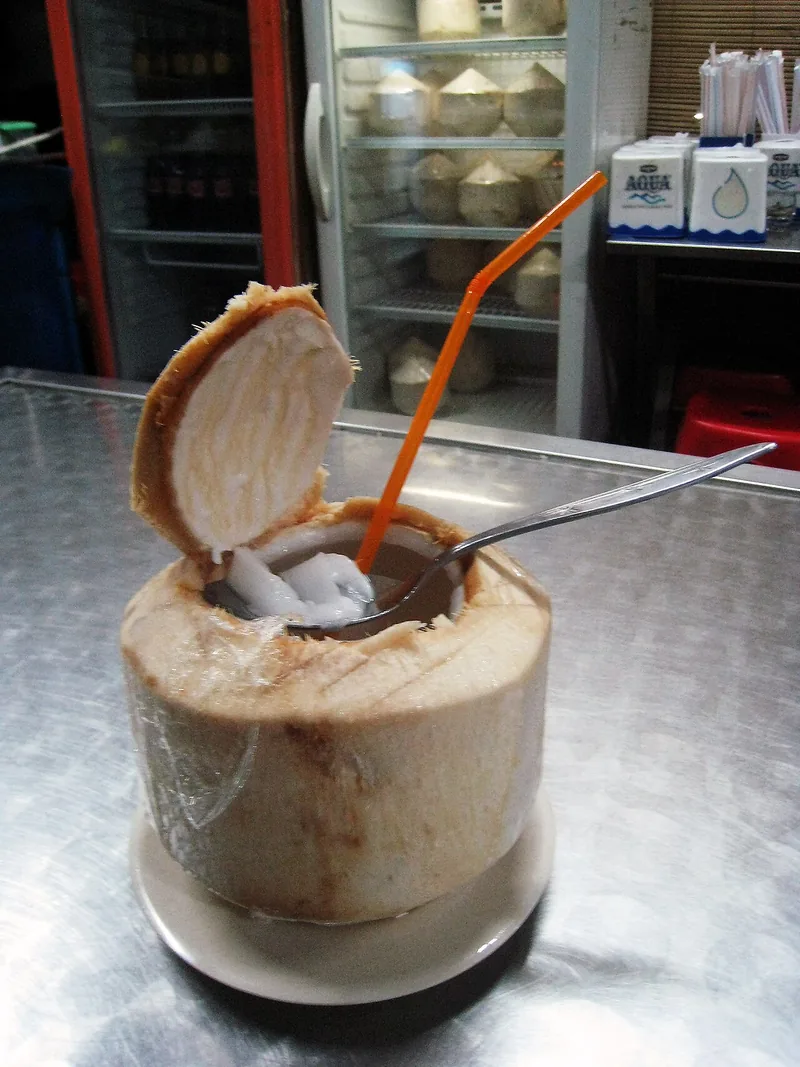 Es kelapa muda disajikan langsung dalam buah kelapa dengan es batu, sendok, dan sedotan oranye