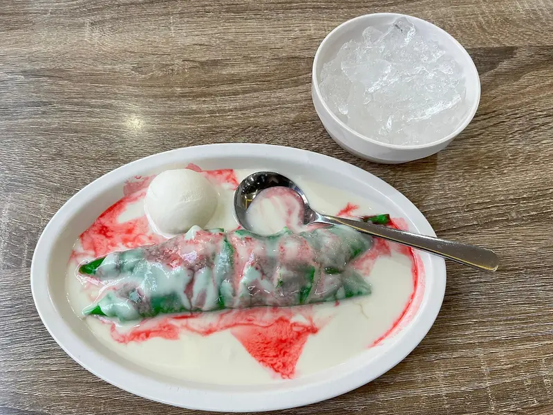 Pisang ijo utuh di piring oval, disiram bubur sumsum putih dan sirup merah, dengan mangkuk es batu