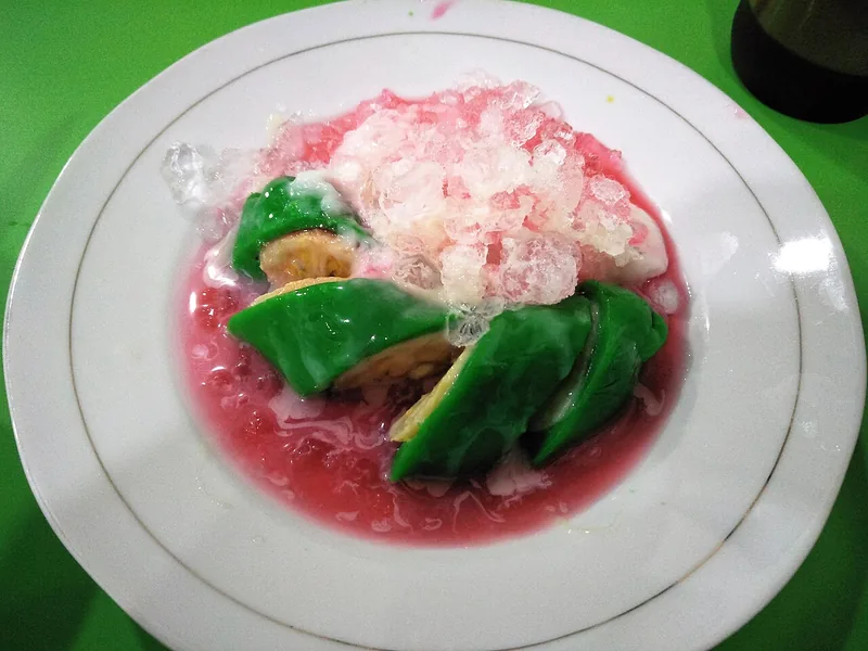 Potongan pisang ijo dengan lapisan adonan hijau tebal membungkus pisang, dalam kuah sirup merah dan es serut