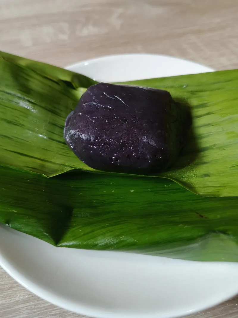 Sepotong kue bugis berwarna ungu tua disajikan di atas daun pisang dalam piring putih