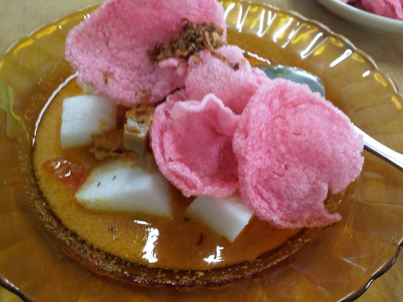 Potongan lontong putih dalam kuah santan kekuningan dengan kerupuk merah muda di piring kaca
