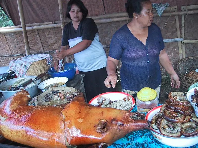 Babi guling utuh berkulit cokelat mengilap di meja penjual bersama mangkuk berisi sosis dan lauk lain