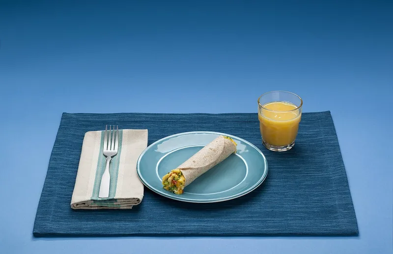Piring berisi roti gulung isi telur dan sayur dengan segelas jus jeruk di atas alas biru