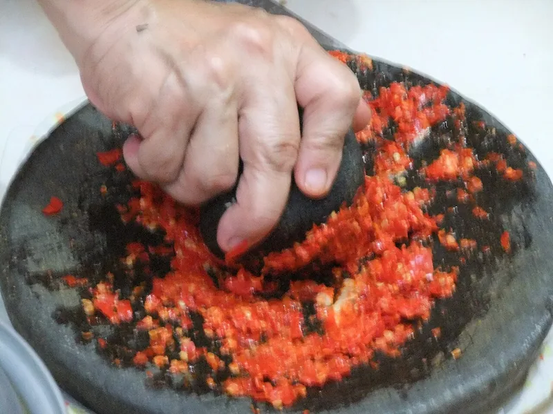 Tangan mengulek cabai merah di cobek batu hingga menjadi bumbu cabai halus