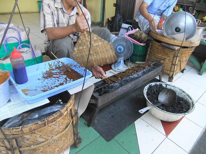 Pedagang membakar sate daging kerbau di atas pemanggang arang sambil mengipas dengan kipas anyaman bambu