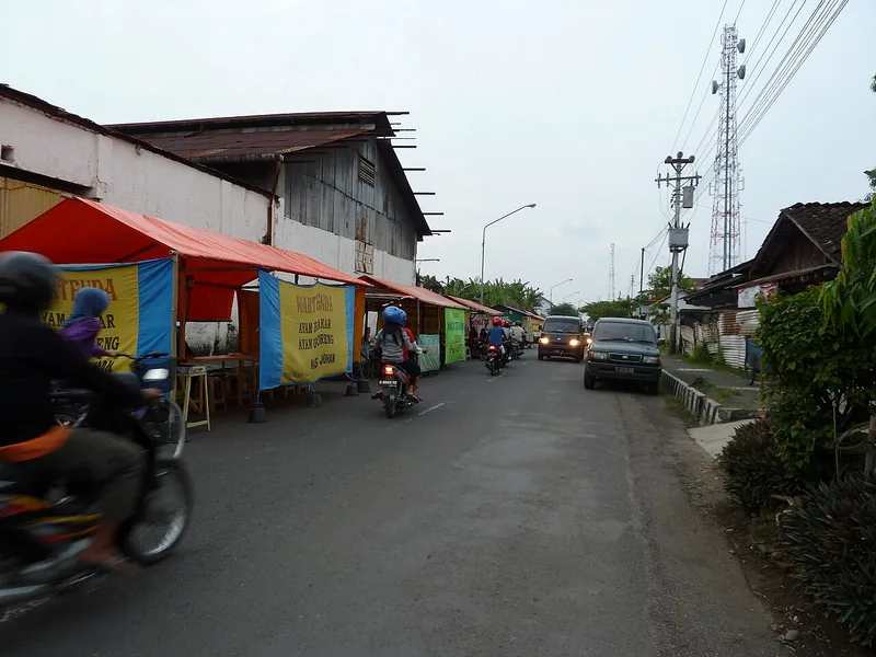 Deretan warung tenda berspanduk di tepi jalan dengan sepeda motor dan mobil melintas