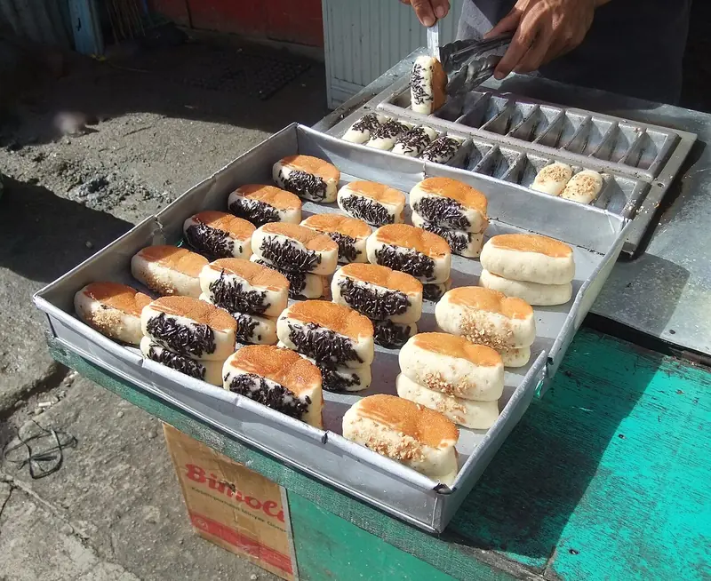 Deretan kue pancong berlapis meses cokelat dan kacang di nampan pedagang dengan cetakan logam