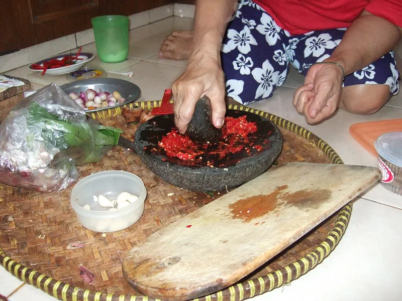 Tangan mengulek cabai merah di cobek batu dengan ulekan, bawang putih dan bawang merah di sampingnya