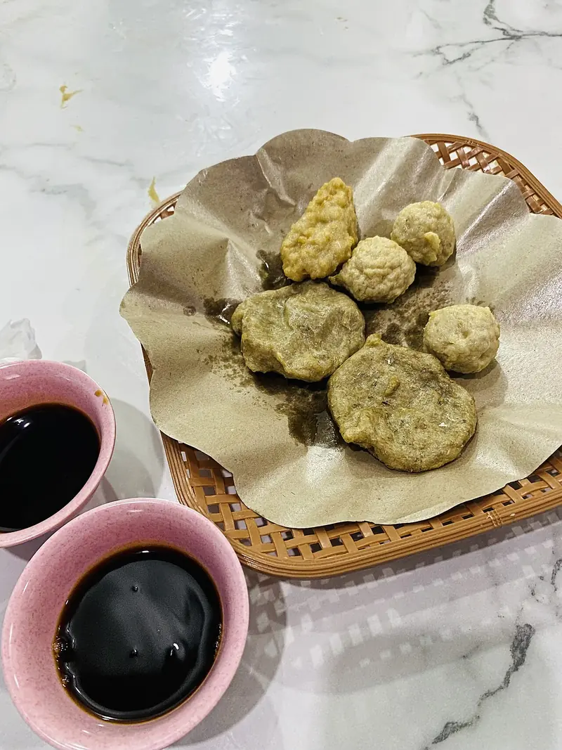 Dua mangkuk kecil berisi kuah cuko hitam di samping pempek goreng di atas keranjang anyaman