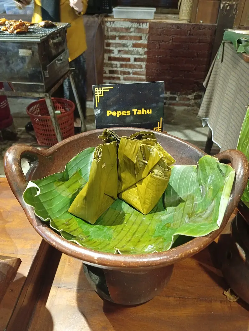 Dua bungkus pepes tahu terlipat rapi dengan daun pisang hijau di dalam wadah tanah liat bergagang