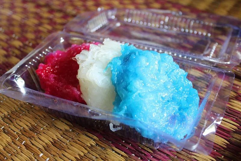 Putu mayang warna merah muda, putih, dan biru dalam kotak mika di atas tikar anyaman