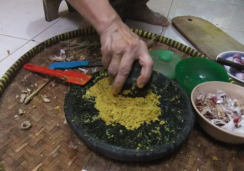 Tangan mengulek bumbu kunyit berwarna kuning di cobek batu hitam di atas tampah anyaman