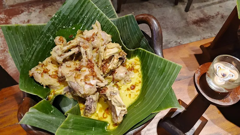 Opor ayam kampung bertabur bawang goreng disajikan di atas daun pisang dalam wadah tanah liat