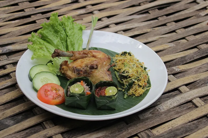 Ayam goreng dengan sambal matah, sayur urap, selada, timun, dan tomat di piring beralas daun pisang