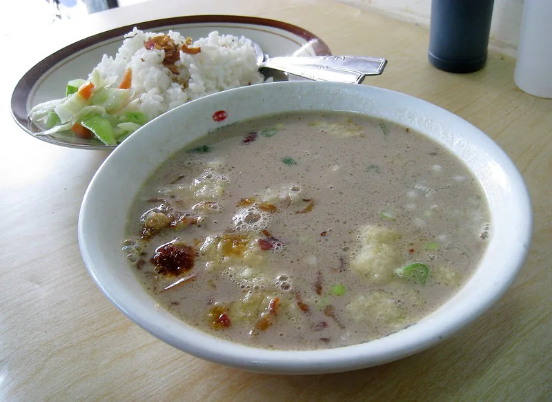 Semangkuk soto betawi berkuah santan pucat dengan potongan daging, di samping sepiring nasi dan acar