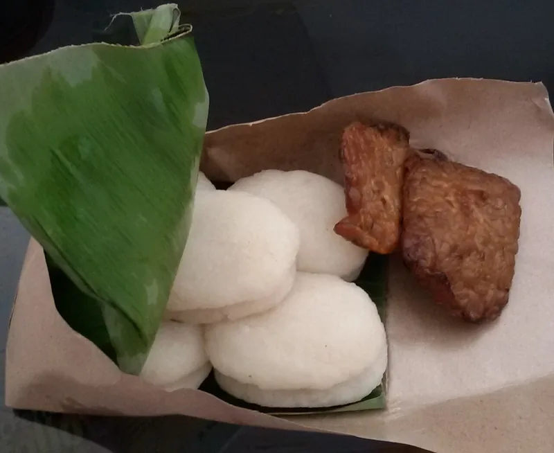 Dua potong tempe bacem cokelat disajikan bersama jadah ketan putih beralas daun pisang