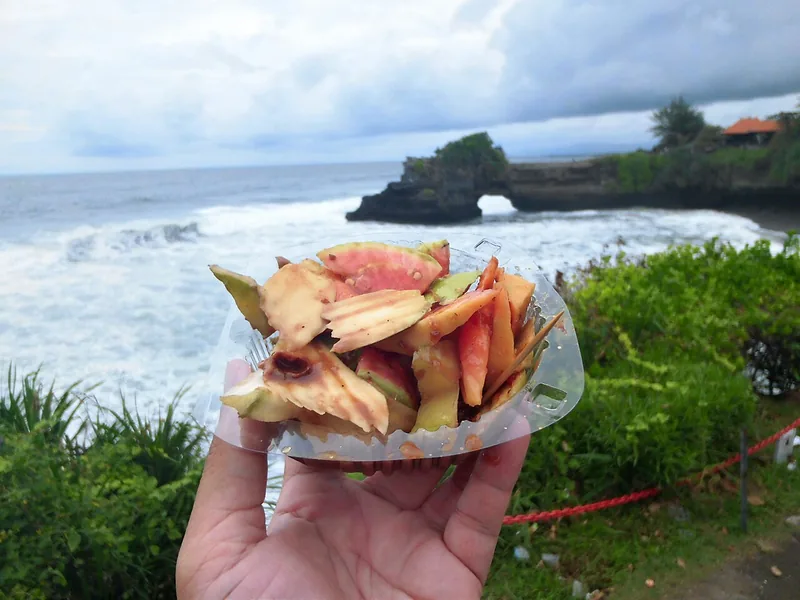 Rujak buah dalam wadah plastik dipegang tangan dengan latar laut dan tebing karang