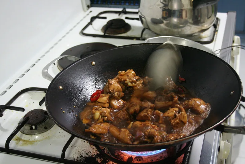 Potongan ayam dimasak dengan kuah kecap yang mulai menyusut di wajan di atas kompor gas