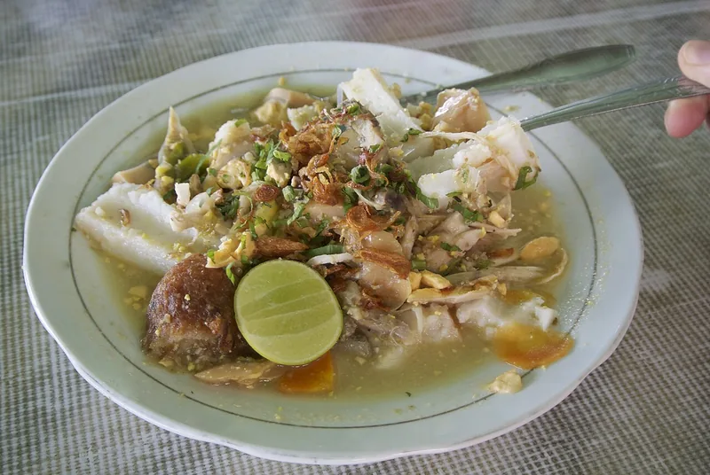 Soto Banjar dengan potongan ketupat, suwiran ayam, bawang goreng, daun bawang, dan jeruk nipis di piring