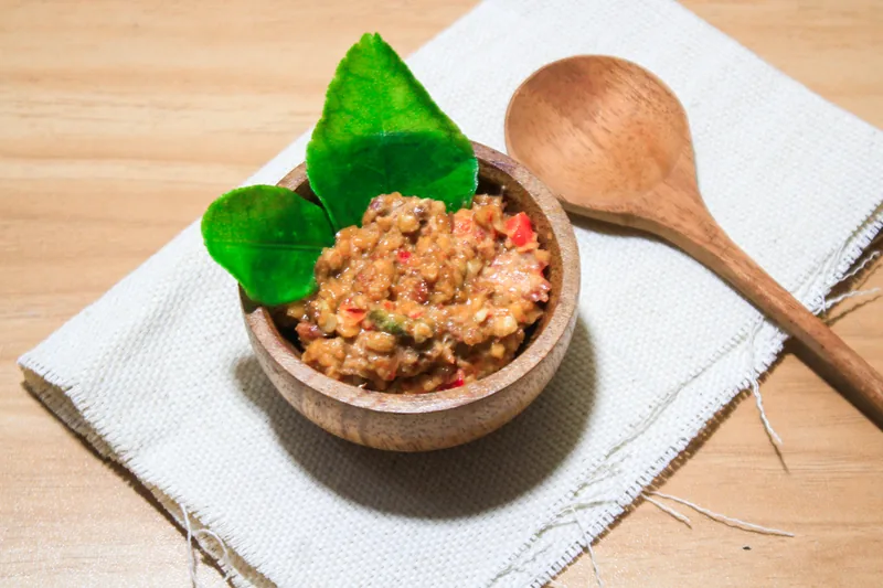 Sambal kacang bertekstur kasar dengan potongan cabai merah dalam mangkuk kayu dan dua daun jeruk