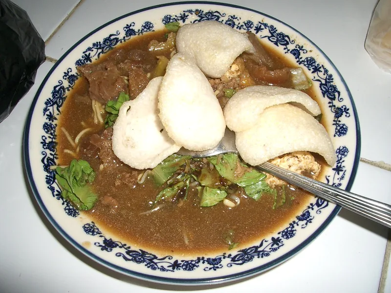 Tahu campur berkuah petis cokelat dengan daging, sayuran hijau, mi, dan kerupuk di piring bermotif biru