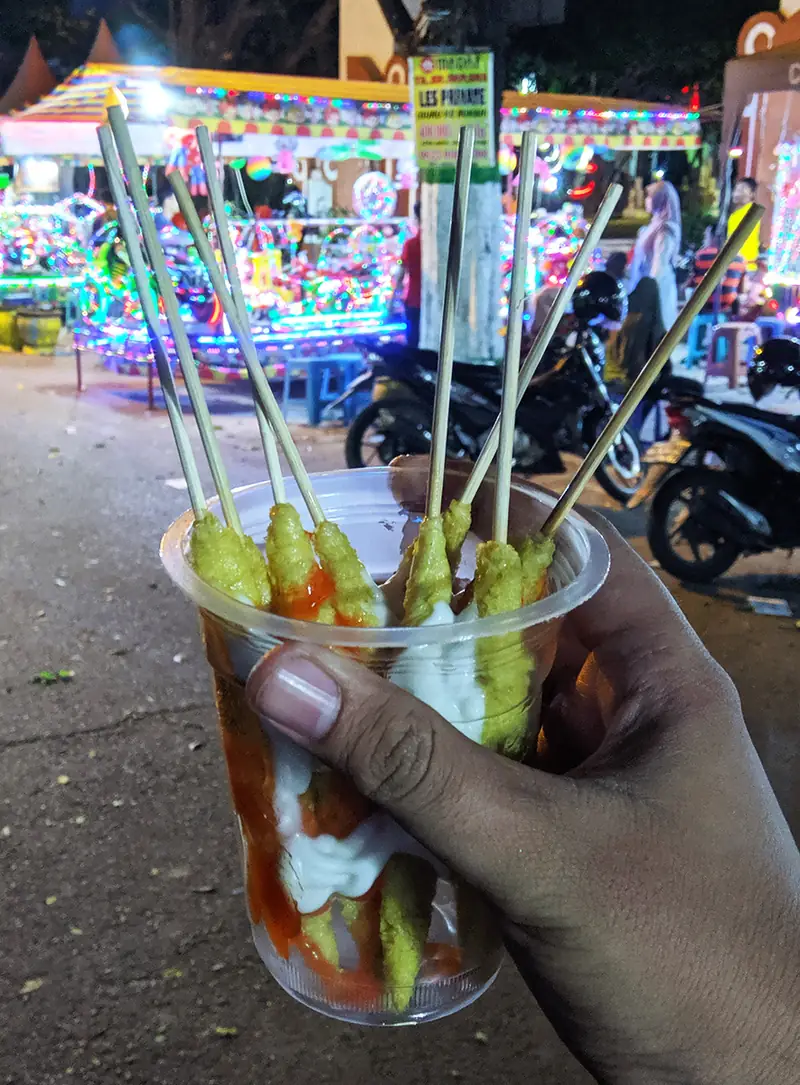 Tangan memegang gelas plastik berisi sate telur gulung bersaus sambal dan mayones di alun-alun malam hari