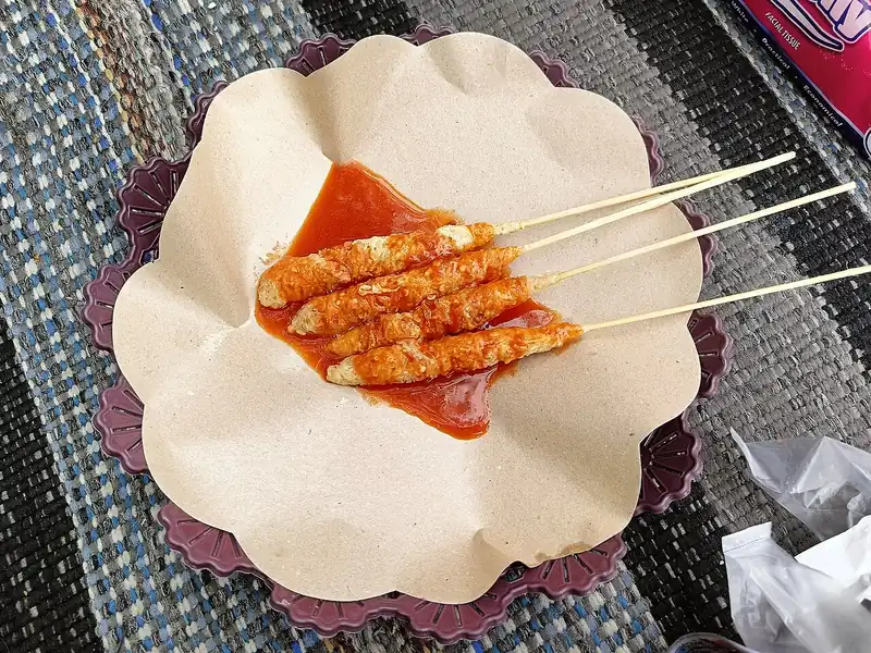 Empat tusuk telur gulung isi bihun disiram saus sambal merah di atas kertas nasi