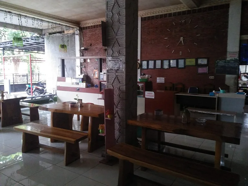 Ruang makan warung dengan meja dan bangku kayu panjang, dinding bata merah, dan jam dinding