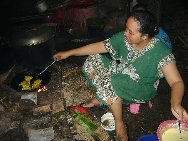 Seorang ibu menggoreng tempe mendoan di wajan di atas tungku kayu dengan adonan tepung di sampingnya