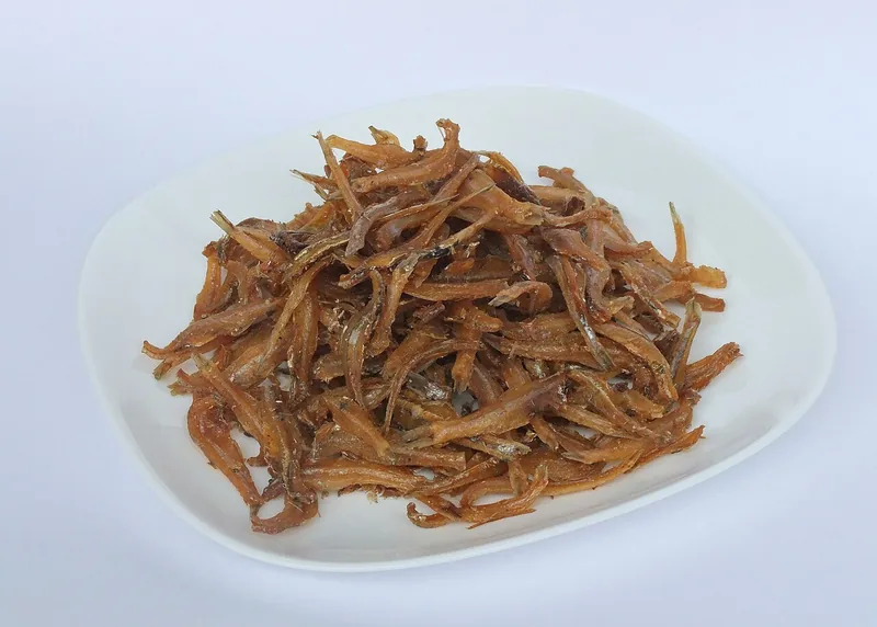 Ikan teri goreng kering berwarna cokelat keemasan ditumpuk di atas piring putih