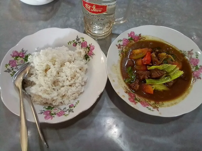 Sepiring nasi putih dan semangkuk tongseng kambing berkuah gelap dengan kol dan tomat di meja